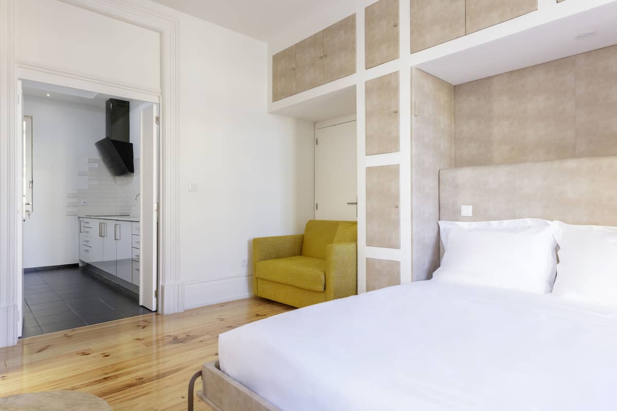 A bedroom with a bed, a yellow armchair, and a view into the kitchen.