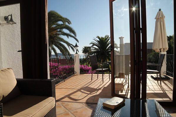 A balcony with a wicker sofa, a table with a book, and a view of palm trees and flowers. - Luxury Detach Villa, Air Con, Private Pool with sun all day, Stunning Sea Views. (Teulada)