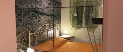 Un dormitorio con cama, mesita de noche, lámpara, espejo y ventana con cortinas.