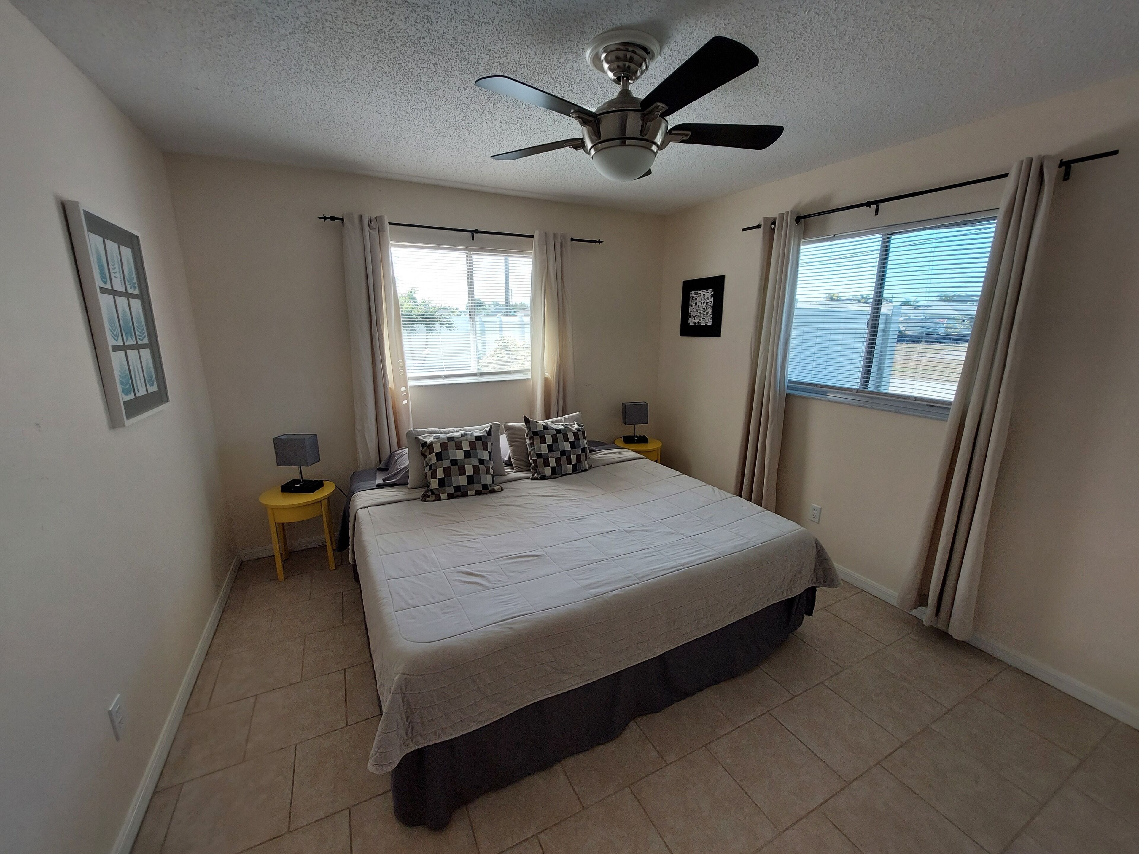 A bedroom with a bed, a ceiling fan, two bedside tables with lamps, and windows with curtains.