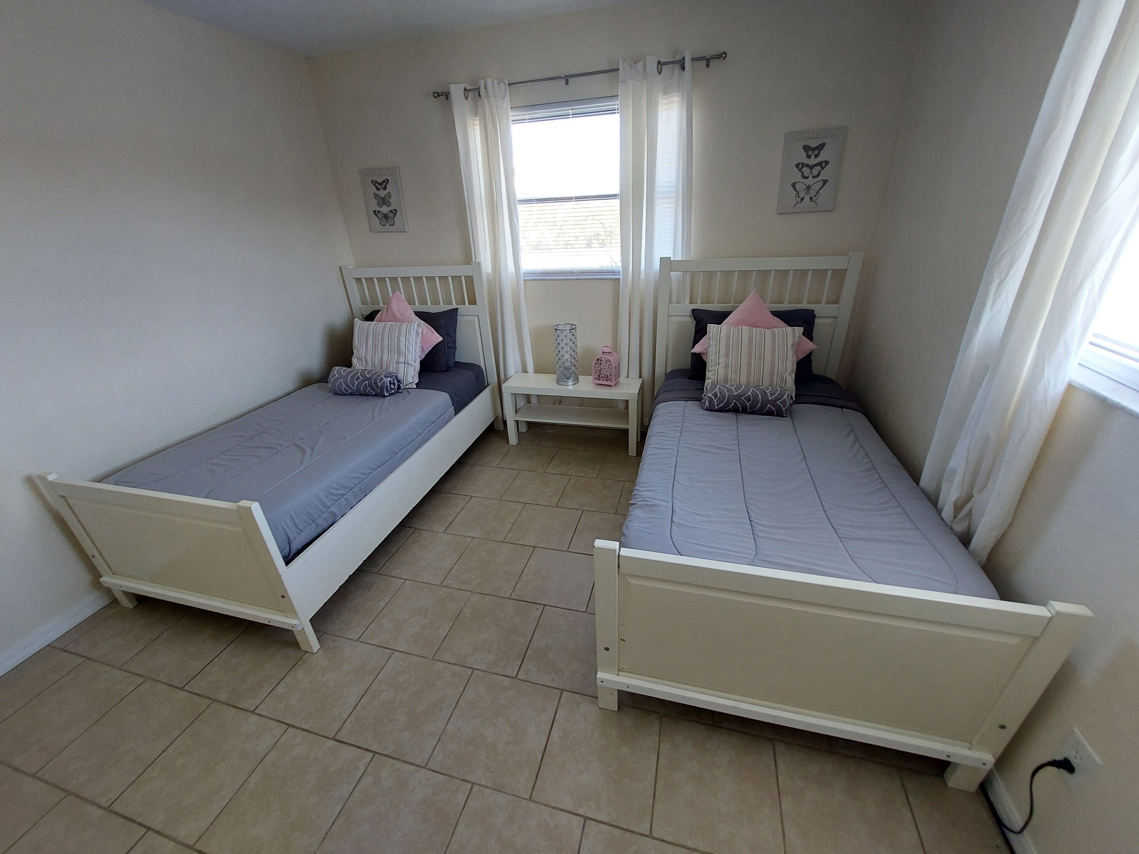 Een slaapkamer met twee eenpersoonsbedden, een klein nachtkastje, een raam met gordijnen en vlinderdecoratie aan de muur.