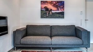 A modern living room with a grey sofa, a mounted TV, and a painting of a moose on a hill. - Vacation apartment Hochgrat (Vogt)