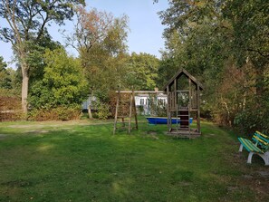 Une cabane en bois, avec une échelle et une balançoire, se trouve dans un jardin verdoyant, parsemé d’arbres, et un bateau bleu est visible au loin.