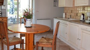 Une table à manger en bois avec des chaises, un coin cuisine avec des meubles blancs et une vue sur l’extérieur grâce à une porte vitrée.