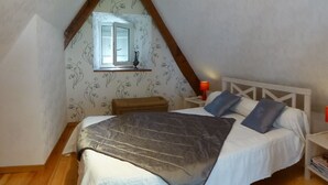 Une chambre confortable avec un plafond en pente, un lit recouvert d’une couverture grise, deux oreillers, une table de chevet avec une lampe rouge et une petite fenêtre avec un rideau.