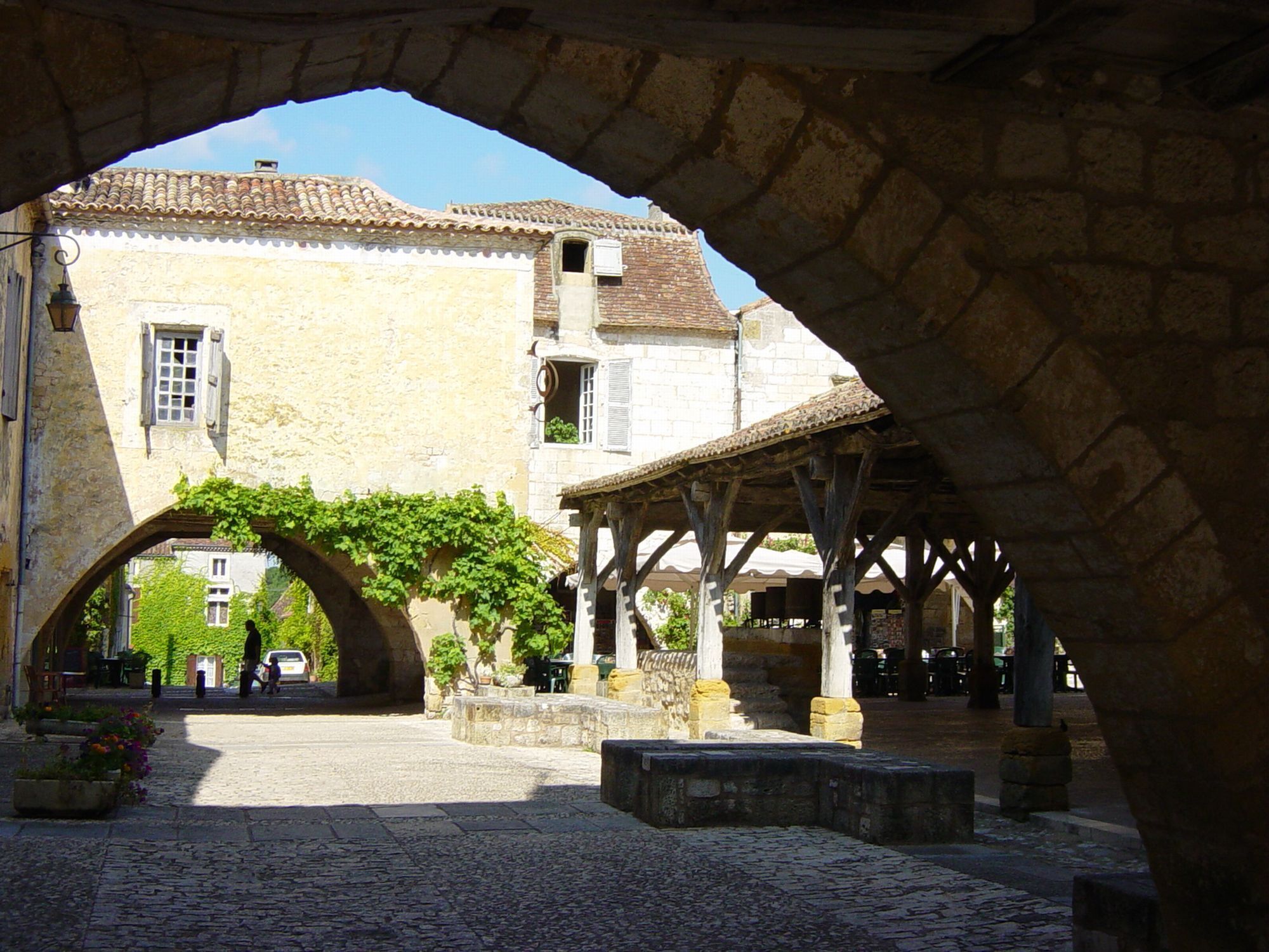A stone archway leading to a courtyard with cobblestone pavement, a covered seating area, and a building with a tiled roof and windows.