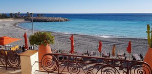 Une terrasse en bord de mer, avec des tables et des chaises, une rambarde et une vue sur la plage et l’océan.