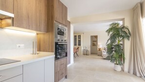 Une cuisine moderne avec des meubles en bois, un four encastré et un plan de travail. On y trouve un coin repas avec une table et des chaises, ainsi qu’une grande plante en pot.