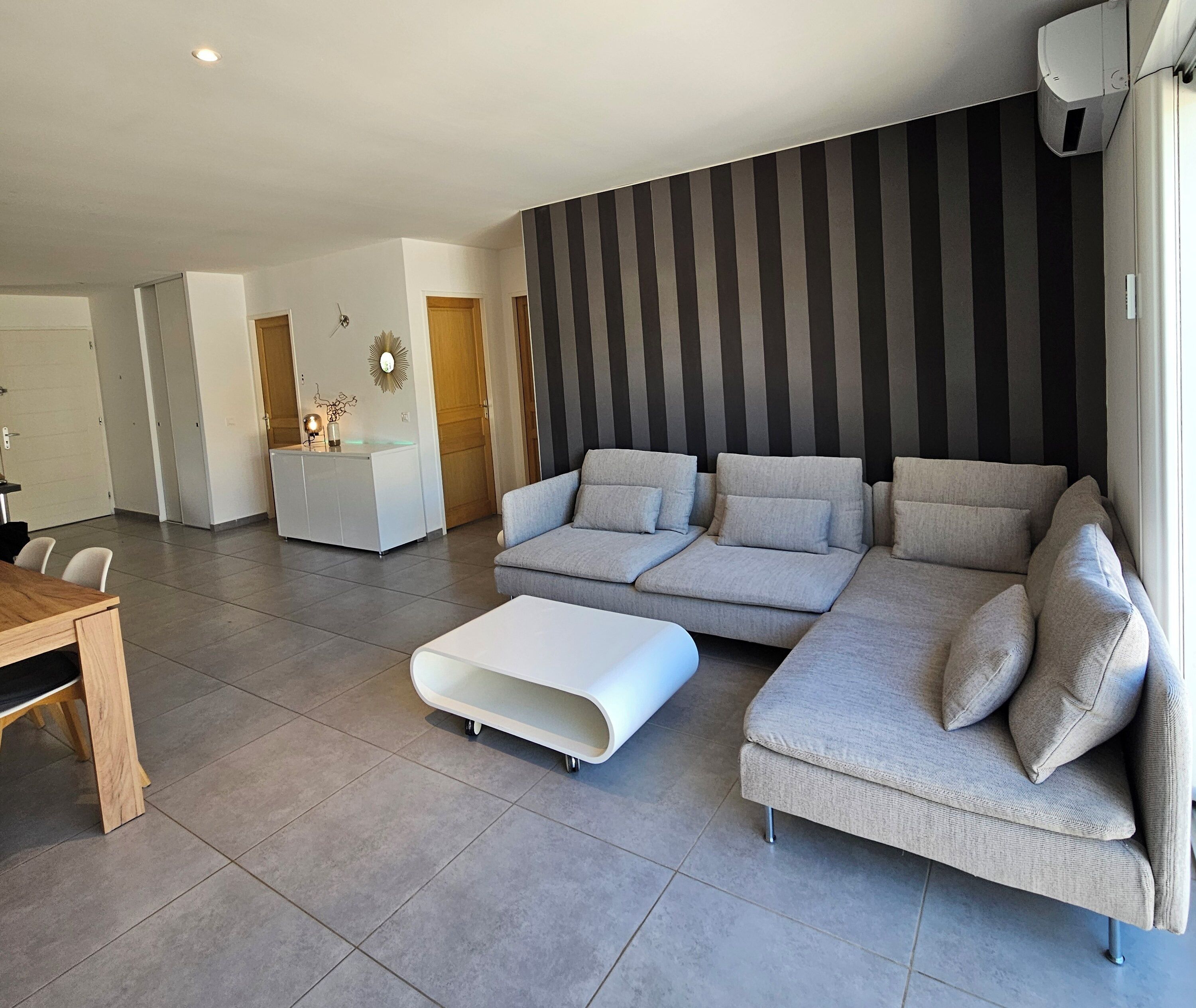 A modern living room with a sectional sofa, a white ottoman, and a kitchen area in the background.
