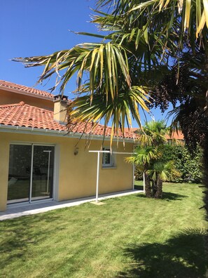 Une maison avec un toit recouvert de tuiles rouges, une porte-fenêtre coulissante et un petit jardin agrémenté d’un palmier.