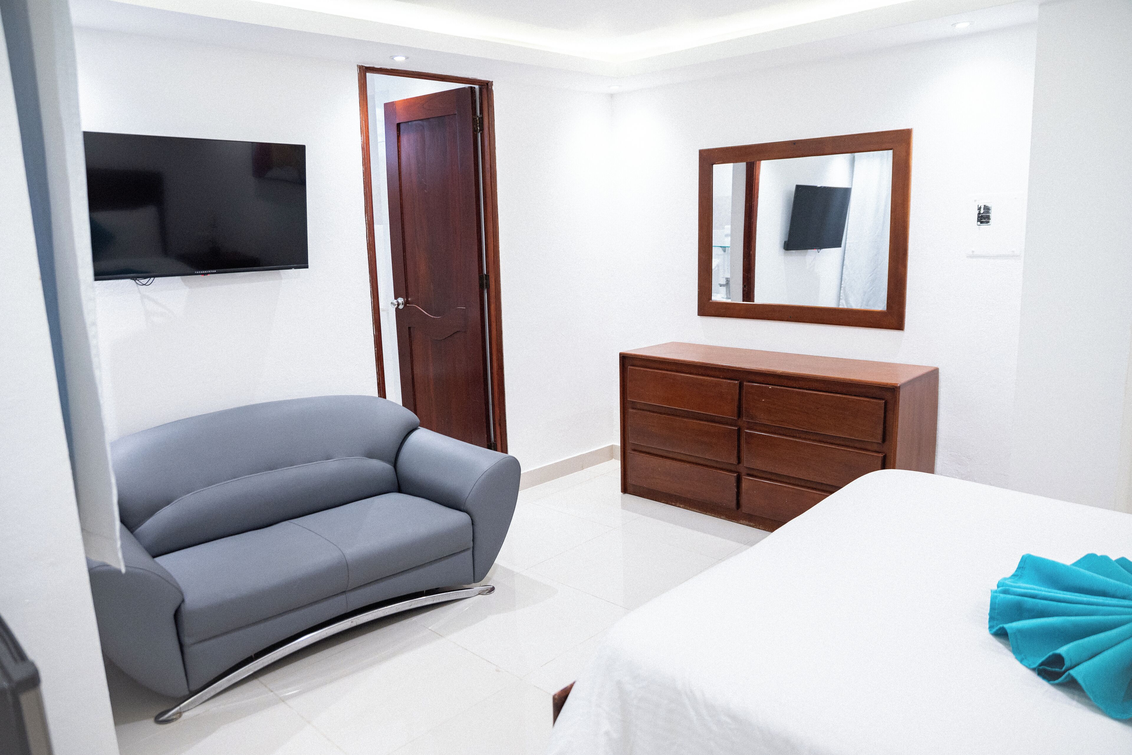 A modern hotel room with a grey sofa, a wooden dresser, a flat-screen TV mounted on the wall, and a bed with a white bedspread.