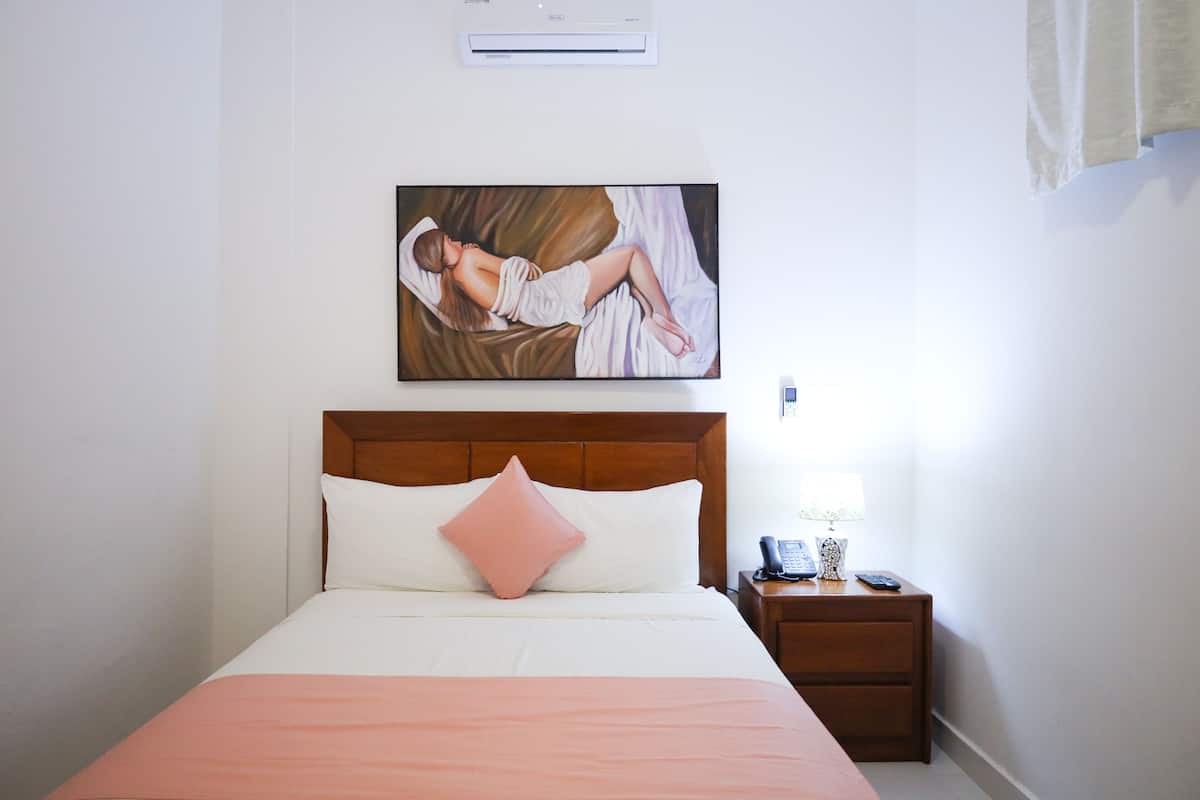A neatly made bed with a pink pillow, a wooden headboard, a bedside table with a lamp, and a painting of a woman on the wall.