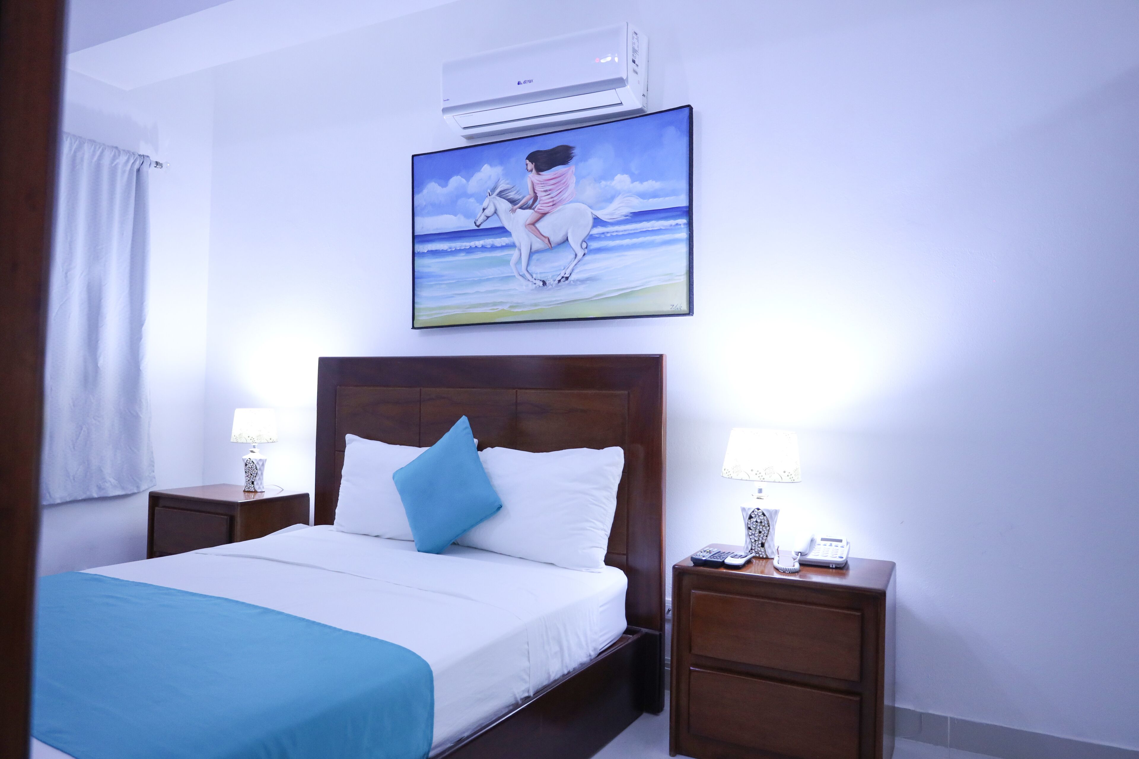 A hotel room with a bed, two bedside tables with lamps, a wall-mounted air conditioner, and a painting of a woman on a horse.