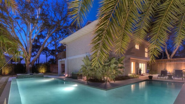 Une piscine rectangulaire, bordée d’une terrasse carrelée, entourée de palmiers et d’un bâtiment dont l’entrée est illuminée.