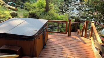 A wooden deck with a hot tub and a view of a forested area with a stream.