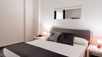 Une chambre d’hôtel bien rangée, avec un grand lit, des tables de chevet, une lampe de chevet, un miroir et un petit objet rouge posé sur l’une des tables.