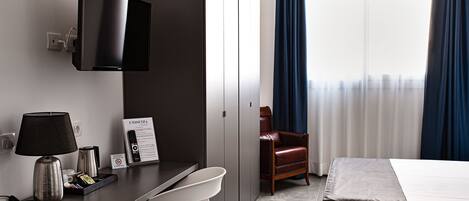 A hotel room with a bed, a desk with a lamp, a chair, a TV mounted on the wall, and a window with curtains.