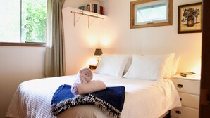 A neatly made bed with a rolled towel and a blanket, a bedside table with a lamp, a shelf with books, and a window with curtains.