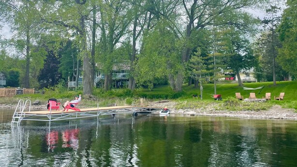 A lakeside view with a dock, chairs, and a dog near the water.