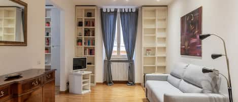 A modern living room with a wooden cabinet, a grey sofa, a TV on a white stand, and bookshelves.