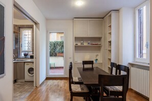 A dining area with a wooden table and chairs, a kitchen with a washing machine, and a view of a garden through a glass door.