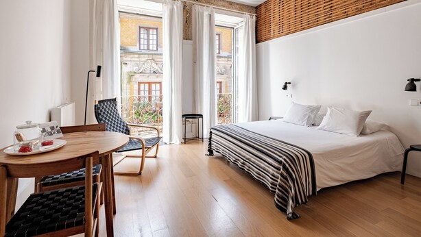 A hotel room with a bed, a dining table with chairs, a small balcony with a chair, and a view of the outside building. - Belomonte Apartments (Porto)