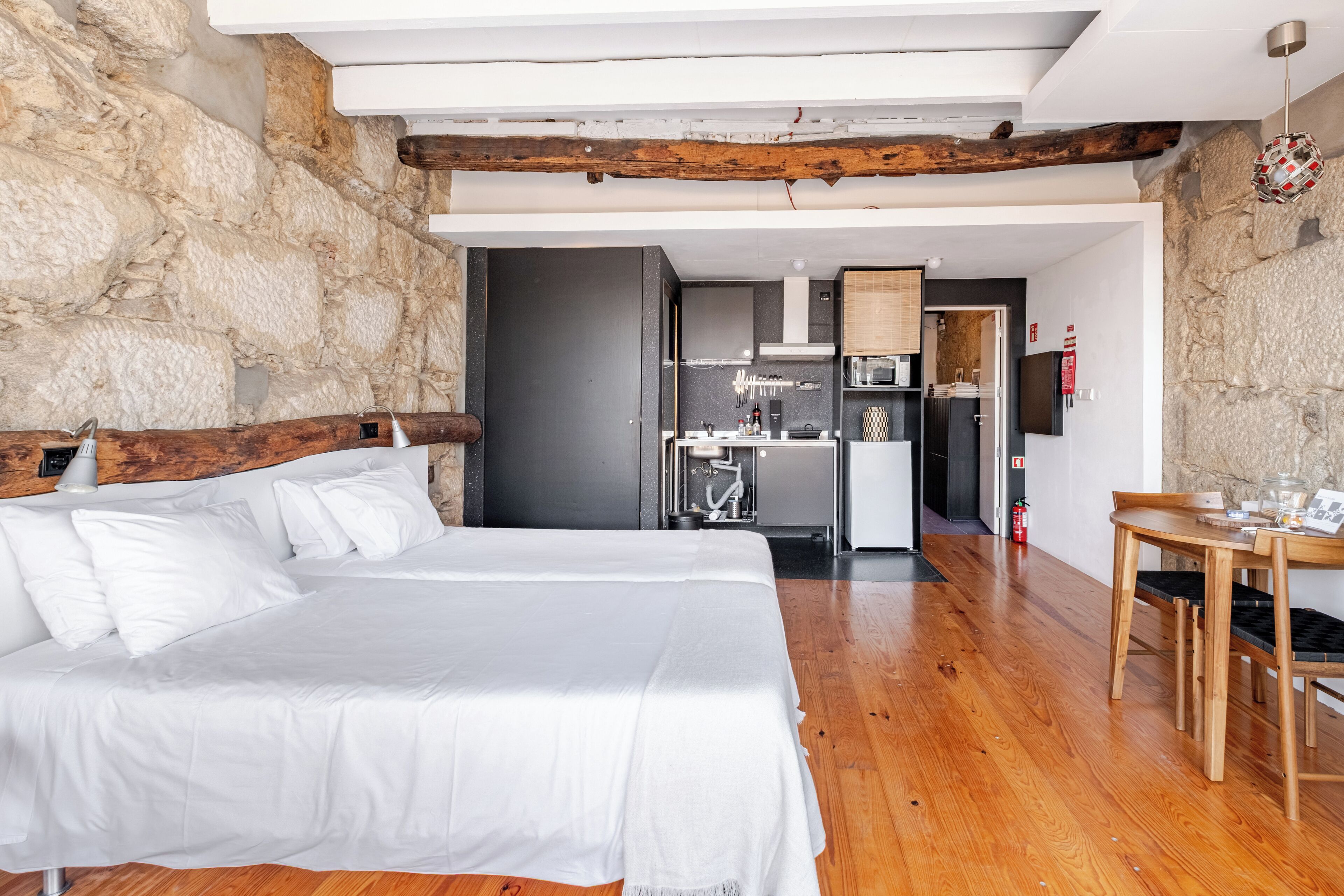 Quarto com cama grande, piso de madeira, paredes de pedra e área de cozinha visível através de uma porta aberta.
