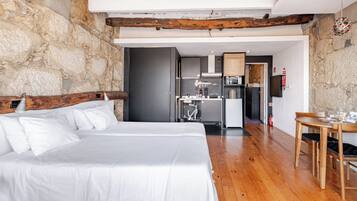 Quarto com cama grande, piso de madeira, paredes de pedra e área de cozinha visível através de uma porta aberta.