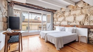A bedroom with a stone wall, wooden floor, two beds, a TV, and a balcony with a view of buildings. - Belomonte Apartments (Porto)