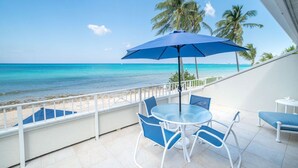 A balcony with a table and chairs, overlooking a beach with palm trees and turquoise water. - 3 Bedroom/2 Bath Oceanfront Condo (Grand Cayman)