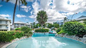 A resort pool area with a clear blue pool, palm trees, and resort buildings in the background. - 3 Bedroom/2 Bath Oceanfront Condo (Grand Cayman)