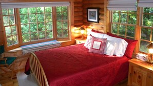 A wooden cabin bedroom with a large bed, a wooden nightstand, a bench, and a window with a view of greenery.