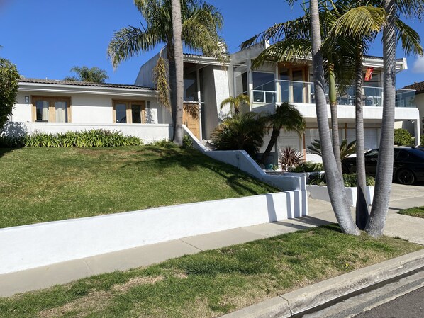 Une maison moderne à deux étages, avec une façade blanche, une pelouse et des palmiers dans le jardin, et une voiture noire garée dans l’allée.