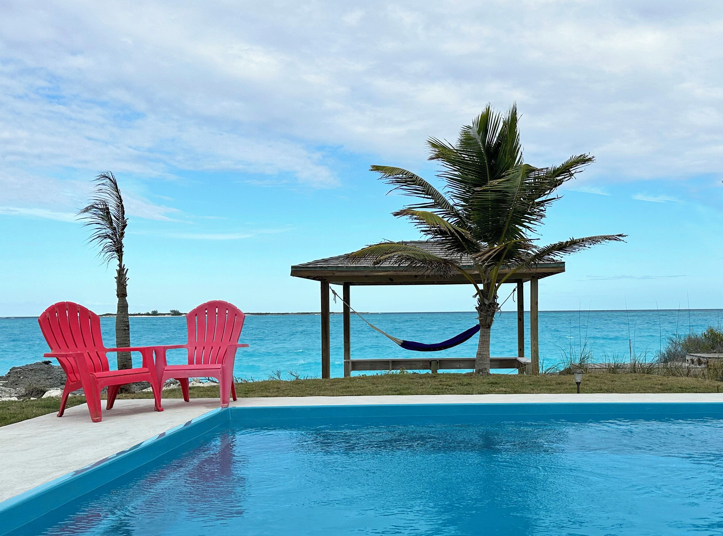 Une piscine avec deux chaises rouges et un hamac, un petit kiosque et une vue sur l’océan.