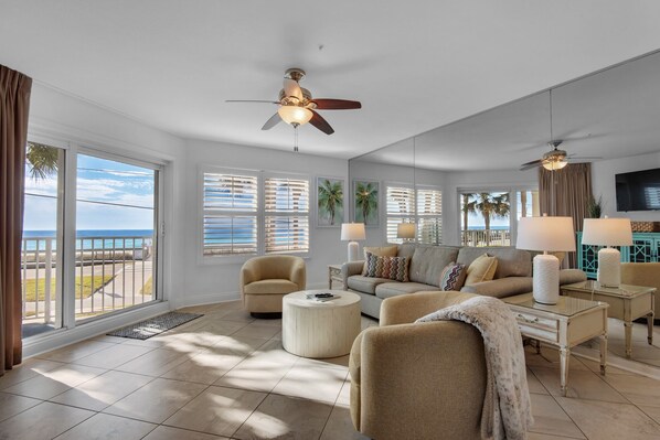 A spacious living room with a large window overlooking the ocean, a ceiling fan, and a television.