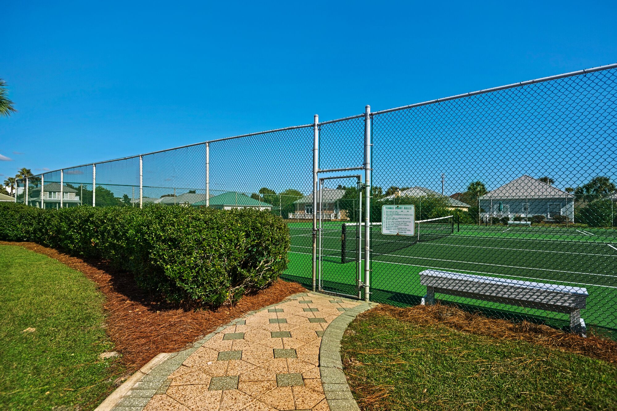 A tennis court with a chain-link fence, a paved walkway, and a bench.