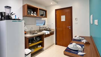 Cozinha compacta com geladeira, micro-ondas e cafeteira. Há um armário de madeira acima da pia e uma mesa de jantar posta com pratos e copos.