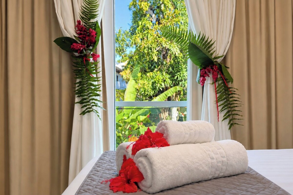 A hotel room with a bed, beige curtains, and a window with a view of greenery.