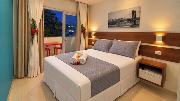 Habitación de hotel con cama, mesitas de noche, cortinas y vistas a la ciudad.