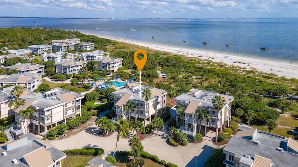 A coastal residential area with multiple two-story buildings, a swimming pool, and a beach in the background. - Amazing Direct Ocean Gulf Front Beach Views Condo, pool and hot tub are open! (Sanibel)