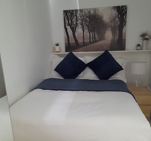 Una cama bien tendida con ropa de cama blanca y azul marino, una almohada azul oscuro y un cuadro enmarcado encima.