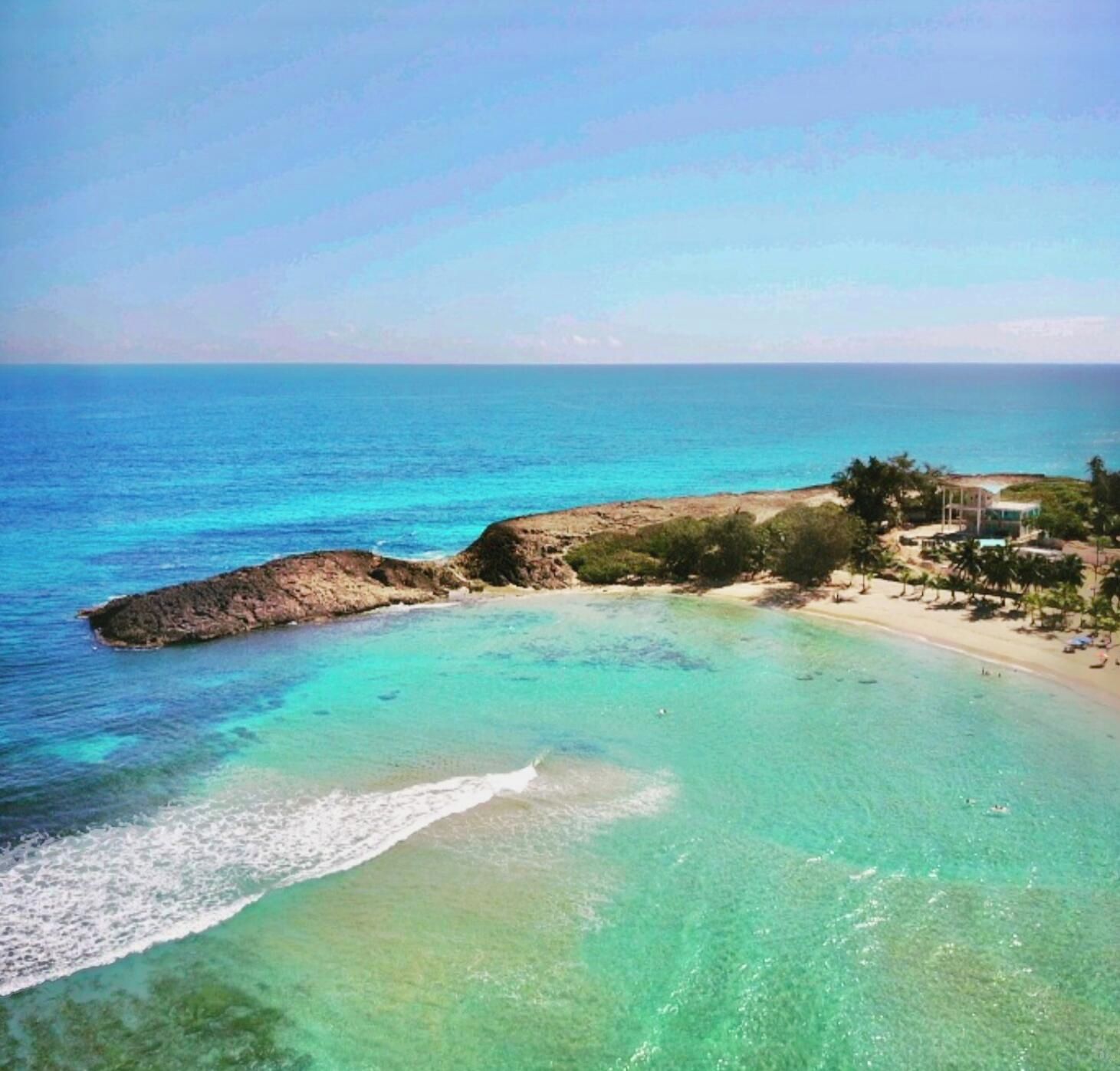 Vista costera con aguas turquesas cristalinas, un promontorio rocoso y una playa con palmeras y un edificio.