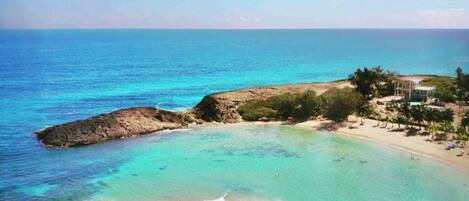 Vista costera con aguas turquesas cristalinas, un promontorio rocoso y una playa con palmeras y un edificio.
