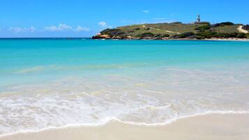 Une plage à l’eau turquoise et limpide, avec un phare qui se dessine à l’horizon.