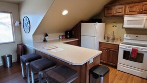 Una cocina con una isla central, armarios de madera, un refrigerador blanco, un microondas y una estufa. Hay tres taburetes en la isla, un bote de basura y un reloj en la pared.