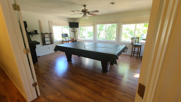 A room with a pool table, wooden flooring, a ceiling fan, and large windows with a view of greenery outside. - Lakefront On A Quiet Cove At Claytor Lake. 
Spring and Summer dates nearly gone! (Dublin)
