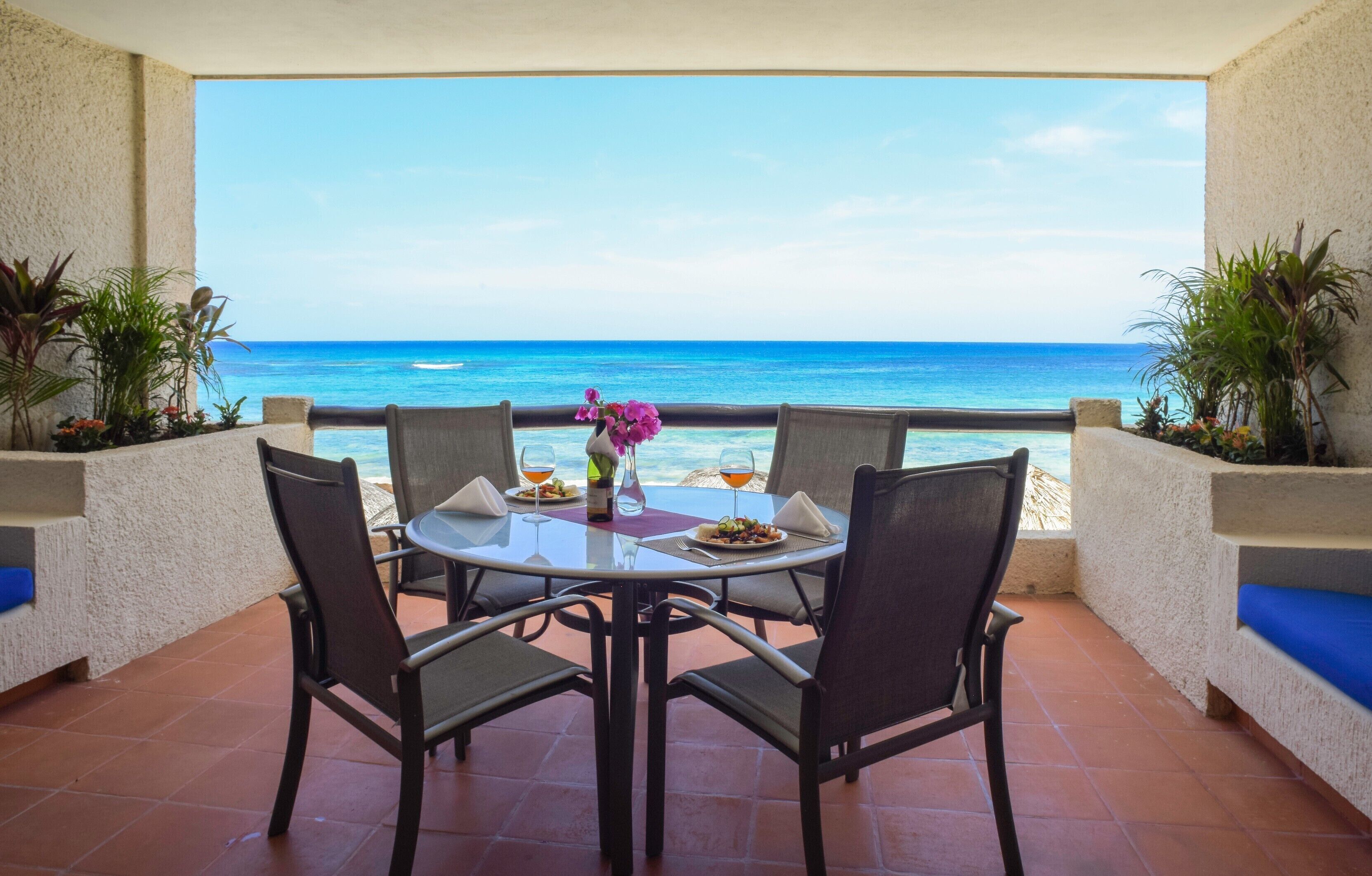 A table with four chairs, a vase with flowers, and a bottle of wine on a terrace with a view of the ocean.