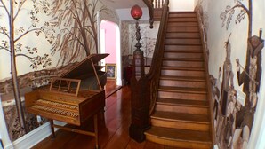 Uma escadaria com degraus de madeira e corrimão, que leva a um patamar com um piano e um mural de árvore na parede.