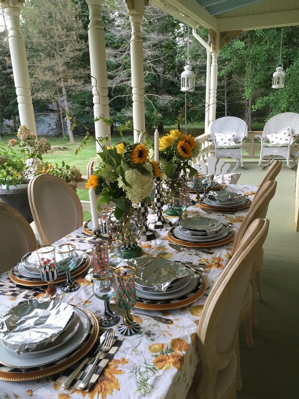 Un espacio exterior techado para comer, con una mesa lista para una comida, sillas alrededor y decoración de flores y velas.