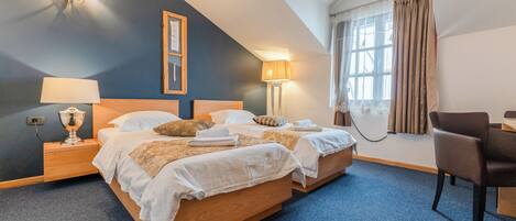 A hotel room with two beds, a nightstand with a lamp, a chair, a window with curtains, and a wall-mounted lamp.